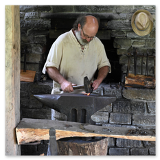 Ein Schmied bearbeitet glühendes Metall auf einem Amboss im Rahmen des mittelalterlichen Burgbauprojekts in Friesach. Umgeben von Steinwänden, Holzträgern und traditionellen Werkzeugen entsteht eine authentische Werkstattatmosphäre. Ein Strohhut hängt an der Wand, das Feuer lodert im Hintergrund – ein Moment voller Konzentration, Kraft und historischer Handwerkskunst.