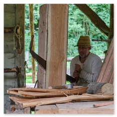 Ein Handwerker bearbeitet mit einer großen Säge einen senkrecht eingespannten Holzstamm in einer offenen Werkhütte beim Burgbauprojekt in Friesach. Er trägt einen Strohhut und ein langärmeliges Hemd, umgeben von frisch geschnittenen Holzplanken und traditionellen Werkzeugen. Die Szene zeigt konzentriertes Arbeiten mit einfachen Mitteln und vermittelt die Atmosphäre mittelalterlicher Baukunst inmitten der Natur.