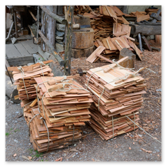 Mehrere Bündel von Holzschindeln liegen sorgfältig mit Seil verschnürt auf einem Erd- und Kiesboden vor einer Werkhütte beim Burgbauprojekt in Friesach. Im Hintergrund sind weitere Holzstücke und Werkzeuge zu sehen, die auf traditionelle Verarbeitung hindeuten. Die Szene vermittelt die Atmosphäre eines mittelalterlichen Handwerksplatzes, an dem mit Geduld und Geschick Materialien für den Burgausbau vorbereitet werden.