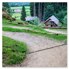 Ein gewundener Pfad führt durch eine grüne Wiesenlandschaft hinauf zu zwei Holzhäusern am Waldrand beim Burgbauprojekt in Friesach. Die Gebäude mit Schindeldächern liegen eingebettet zwischen Bäumen und Hügeln, während im Hintergrund ein rotes Dach und bewaldete Höhen sichtbar werden. Die Szene vermittelt die ruhige Annäherung an den Burgturm – ein Weg durch Natur und Geschichte, der den mittelalterlichen Geist spürbar macht.