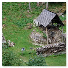 Ein steinerner Ofen mit Holzüberdachung steht eingebettet in eine bewaldete Hanglage beim Burgbauprojekt in Friesach. Daneben lagern gestapelte Holzscheite unter einem offenen Schutzdach, während ein kleiner Weg und ein Hinweisschild zur Anlage führen. Die Szene vermittelt die ruhige Kraft traditioneller Handwerksplätze und zeigt, wie Geschichte durch einfache Materialien und menschliches Können lebendig wird.