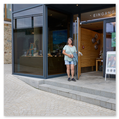 Person mit Jacke vor modernem Eingang mit Glasfront und Steinmauer, Infotafel und Pflanzgefäß im Weingut Taggenbrunn