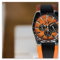Sportlicher Chronograph mit orange-schwarzem Zifferblatt, Metalllünette und passendem Armband, ausgestellt auf weißem Sockel in der Jacques-Lemans-Uhrenschau bei Taggenbrunn