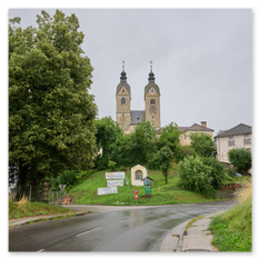 Kirche Maria Saal mit zwei Zwiebeltürmen und goldenen Kugeln, umgeben von Bäumen und Schildern – steinerne Würde auf Anhöhe, bei grauem Himmel und nasser Straße.