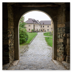 Blick durch alten Steinbogen auf gepflasterten Weg und helle Gebäude mit roten Dächern – gepflegte Wiese und Sträucher rahmen den ruhigen Hofraum bei Taggenbrunn.