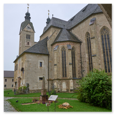 Gotische Kirche Maria Saal mit Turm und Spindeln, gesehen durch Steinbogen – Pflasterweg, Denkmal und Infotafeln im Vordergrund, moosige Mauern und historische Tiefe.