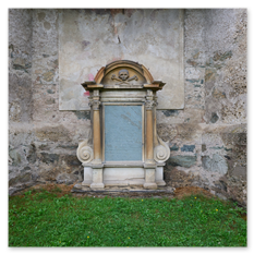 Altes Grabdenkmal mit Totenkopfrelief und verwittertem Schriftfeld vor Steinmauer bei Maria Saal – klassischer Aufbau mit Säulen, Grasfläche und Zeichen der Vergänglichkeit.