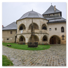Historischer Bau mit Turm und Rundbögen bei Maria Saal – gepflasterter Weg, Denkmal im Gitter und Sitzbereich im Hof, nasse Steine unter grauem Himmel.