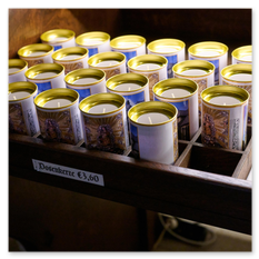 Holzregal mit Dosenkerzen in goldenen Dosen bei Maria Saal – religiöse Bildetiketten, Preisangabe und Broschürenstapel, geordnete Andachtsökonomie im Kirchenraum.