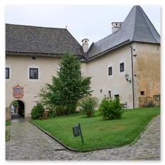 Historisches Torhaus mit Wappen über dem Bogen bei Maria Saal – Schindeldächer, gepflasterter Weg, Bänke und Grünfläche im ruhigen Dorfbild mit architektonischer Würde.
