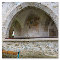 Steinbogen mit verblassten Wandfresken und Bank bei Maria Saal – Sitzplatz vor verwittertem Mauerwerk, religiöse Reste im Bogenraum, Dorfarchitektur im Hintergrund.