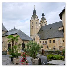 Hofansicht mit Zwiebeltürmen bei Maria Saal – Steinbauten, Freskenreste, Blumentöpfe und Sitzmöbel, gepflasterter Boden mit geparktem Auto unter grauem Himmel.