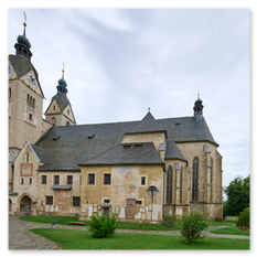 Kirchhof mit Zwiebeltürmen bei Maria Saal – Steinbauten, Freskenreste, Pflanzkübel und Sitzmöbel, gepflasterter Boden mit Auto, sakrale Würde im touristischen Rahmen.