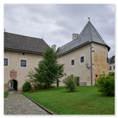 Torbau mit Wappen bei Maria Saal – Schindeldächer, Gasthaus Sandwirt, Grünfläche und Bänke im gepflegten Dorfgefüge, historischer Steinweg unter grauem Himmel.