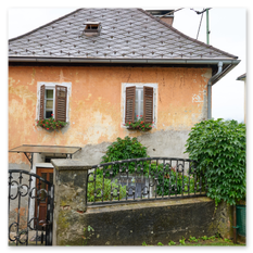 Hausansicht mit orangefarbener Fassade, Fensterläden und Blumenkästen bei Maria Saal – Garten mit Gitterzaun, Busch und Steinmauer im ruhigen Dorfgefüge.