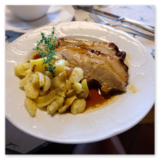 Schweinsbraten mit Bratensaft und Kartoffelbeilage im Gipfelhaus Magdalensberg – angerichtet auf weißem Teller mit Thymianzweig, gedeckter Tisch im Gastraum.