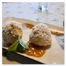 Zwei Marillenknödel mit Staubzucker auf Fruchtsauce im Gipfelhaus Magdalensberg – rechteckiger Teller mit Minze, gedeckter Tisch mit Besteck und Gläsern im Hintergrund.