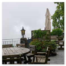 Steinterrasse mit Holzmöbeln und geschlossenen Schirmen am Gipfelhaus Magdalensberg – Nebel beginnt sich zu lichten, Blick ins Tal durch Geländer und Baumkulisse.
