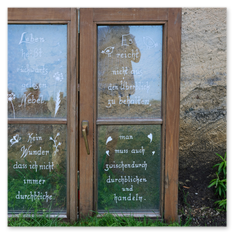 Zwei beschriftete Fensterflügel mit Glasaufschrift im Gras am Magdalensberg – Sprüche über Durchblick und Handeln, Blumenzeichnungen, Steinwand und Pflanze im Hintergrund.