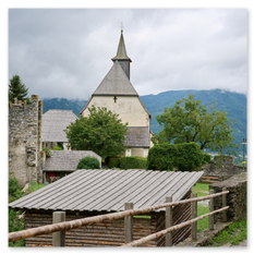 Kirche mit Turm und Burgturm in Friesach – Holzunterstand und Zaun im Vordergrund, Hügellandschaft mit Talblick, Fahnen und Wolkenstimmung im Hintergrund.