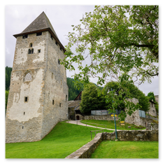 Historischer Burgturm mit Spitzdach in Friesach – Steinmauer mit Fenster, Vorplatz mit Weg und Grünfläche, Waldhang im Hintergrund, ruhige Szene mit Naturanteil.