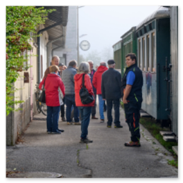 Gruppe von Ausflugsteilnehmern in roten Jacken steht am schmalen Bahnsteig neben dem historischen Zug des Ötscherbahn Express, im Vordergrund ein Bahnmitarbeiter in blauer Uniform, im Hintergrund eine Bahnhofsuhr und grüne Landschaft im leichten Nebel, Moment des Ein- oder Aussteigens