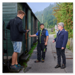Drei Personen stehen neben einem grünen historischen Waggon. Eine Person auf der Einstiegstufe beschäftigt sich mit der Tür, zwei weitere in Uniform beobachten vom Bahnsteig aus, im Hintergrund Steinmauer, Bäume und nebelverhangene Hügel, ländliche und nostalgische Atmosphäre