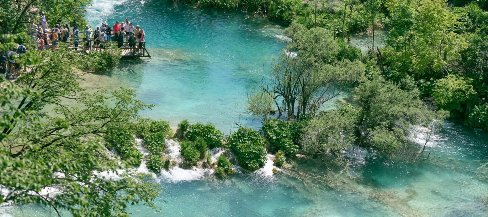 Luftaufnahme einer hölzernen Aussichtsplattform mit Touristen an den Plitvicer Seen mit kristallklarem, türkisblauem Wasser, grünen Bäumen und kleinen Wasserfällen.