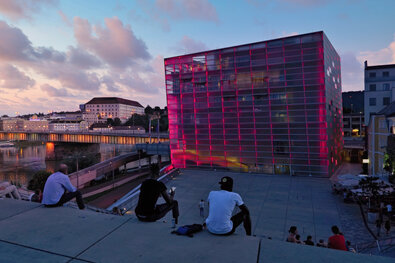 Szene aus Linz mit mehreren Personen, die auf den Treppen vor dem Ars Electronica Center (AEC) sitzen oder stehen. Das moderne Gebäude zeigt seine markante Glasfassade mit pinkfarbiger Beleuchtung und spiegelt den zeitgenössischen Charakter des Ortes wider. Im Hintergrund erhebt sich Schloss Linz auf dem bewaldeten Hügel, wodurch eine visuelle Verbindung zwischen moderner Architektur und historischer Baukunst entsteht. Die Lichtstimmung ist ruhig, vermutlich gegen Abend, wodurch sich warme Reflexe auf dem Wasser und der Fassade zeigen. Die Aufnahme vermittelt eine stille, urbane Lebensszene mit kulturellem Bezug und offenem Stadtraum am Donauufer.