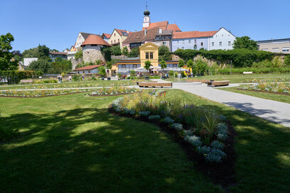 Das Bild zeigt einen stimmungsvollen Blick auf die Orangerie in Schärding, aufgenommen während der Landesgartenschau. Im Vordergrund breiten sich gepflegte Blumenbeete, geschwungene Wege und Sitzbänke aus – umrahmt von üppigem Grün und farbenfroher Blütenpracht. Die Orangerie, ein gelbes Gebäude mit klassischer Architektur, steht zentral und wird vom natürlichen Licht hervorgehoben. Im Hintergrund erkennt man die Altstadt von Schärding, inklusive eines Kirchturms, der charmant aus den umliegenden Gebäuden herausragt. Die Szene verbindet Natur, Architektur und regionale Identität und vermittelt Ruhe und einladende Gartenatmosphäre.