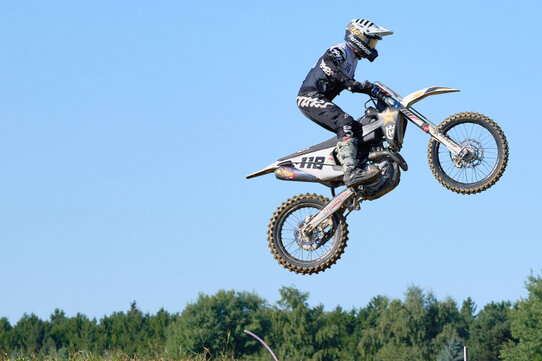 Ein Motocross-Fahrer mit der Startnummer 118 befindet sich in der Luft beim Sprung über eine Erhebung auf der Rennstrecke in Grafenschlag. Der Fahrer trägt vollständige Schutzausrüstung inklusive Helm, Handschuhen und Rennanzug. Das weiße Motorrad hebt sich vor dem klaren blauen Himmel ab, im Hintergrund ist ein dichter Wald mit grünen Bäumen zu sehen. Die Szene zeigt einen präzisen Moment im Rennen und verdeutlicht die Dynamik und Körperbeherrschung im Motocross-Sport.