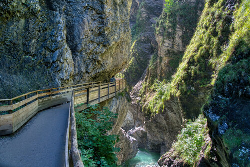 Weg mit Holzgeländer entlang steiler Felswände, darunter türkisfarbener Fluss – ruhige Bewegung durch die Klamm.
