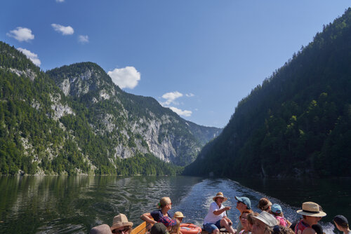 Ein Boot mit mehreren Personen fährt über einen ruhigen See, umgeben von bewaldeten Berghängen. Sommerliches Licht und klare Spiegelungen schaffen eine friedliche Stimmung.
