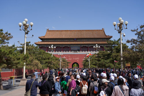 Menschenmenge vor dem südlichen Eingang der Verbotenen Stadt in Peking, dem Meridian-Tor; traditionelle chinesische Architektur mit roter Fassade und verzierten Dachziegeln, flankiert von Bäumen und Straßenlaternen bei klarem Himmel