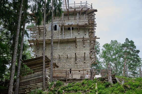 Mittelalterlicher Burgturm im Bau Unvollendeter Turm aus Bruchsteinmauerwerk, umgeben von mehrstöckigem Baugerüst aus Holz. Im Vordergrund Palisade und zusätzliche Holzkonstruktionen. Dahinter hohe Bäume, im Hintergrund teils bewölkter Himmel.