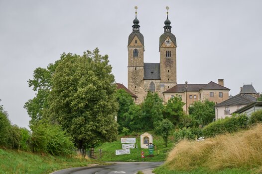 Maria Saal – Doppelturm im Landschaftsbild Kirche Maria Saal mit zwei Zwiebeltürmen und goldenen Kugeln, umgeben von Bäumen und Schildern – steinerne Würde auf Anhöhe, bei grauem Himmel und nasser Straße.