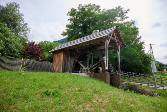 Schaumühle – Kärnten Traditionelle Schaumühle mit Holzrad unter Dachkonstruktion – Wiese, Zaun und Bäume im Umfeld, Wasserlauf daneben, ruhige Szene mit Technikbezug.