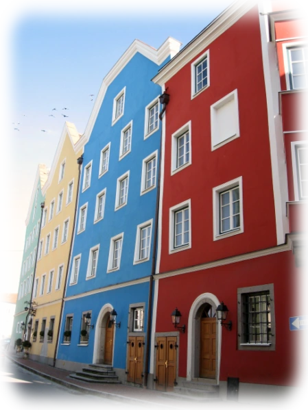 Vier bunt gestrichene Häuser in Schärding, mit klassischer Architektur und dekorativen Fassaden. Das Bild zeigt ein farbenfrohes Häuserensemble in Schärding, bestehend aus vier nebeneinander stehenden Gebäuden in Gelb, Blau, Rot und Beige. Die Fassaden sind liebevoll gestaltet mit dekorativen Fensterrahmen, Holzläden und klaren Linien. Vor den Gebäuden führen kleine Treppen zu hölzernen Eingangstüren, die einladend wirken. Die traditionelle Architektur mit steilen Dächern verleiht dem Straßenzug ein historisches Flair. Die Aufnahme hebt die farbliche Harmonie und den malerischen Charakter der Stadt hervor und lädt dazu ein, die Details der Fassadengestaltung zu entdecken.