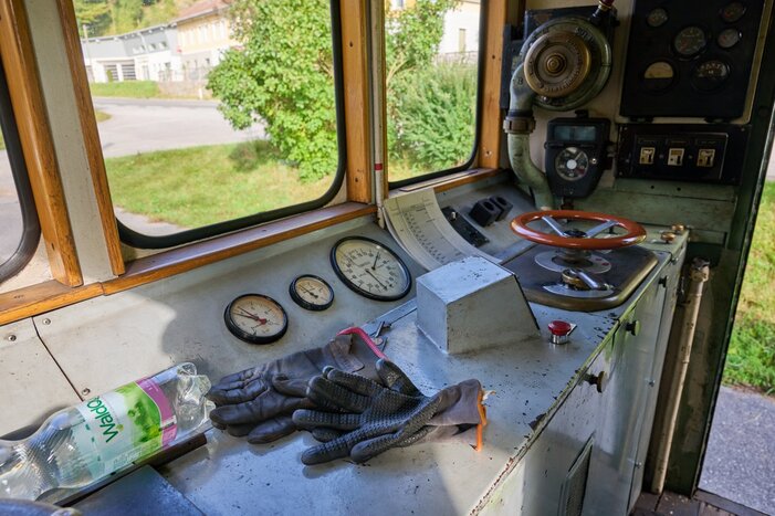 Detailreiches Steuerpult eines älteren Fahrzeugs mit vier Anzeigen, Hebeln, Handschuhen und Vöslauer-Flasche. Blick nach draußen auf Wiese und Gebäude.