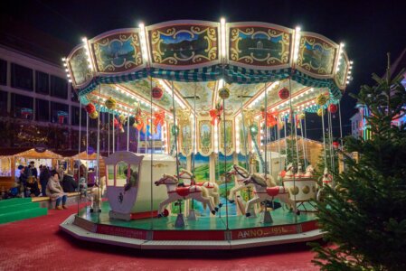 Beleuchtetes Karussell mit weißen Pferden und Kutsche, umgeben von Marktständen und Menschen, rechts ein geschmückter Weihnachtsbaum.