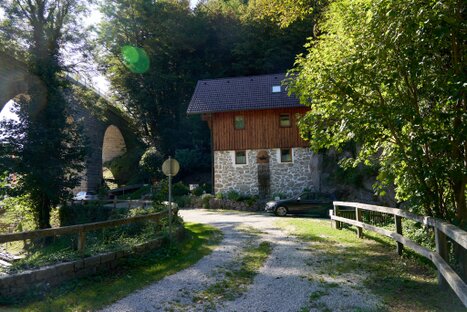 Steinhaus mit Holzaufbau am Waldrand, daneben Viaduktbogen; geschwungene Zufahrt, Sonnenlicht im Grün – Eingang zur Stillensteinklamm, ruhig und einladend.