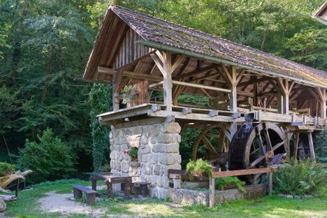 Holzwasserrad auf steinerner Basis, umgeben von Waldgrün; Blumentöpfe, Sitzbank und Lichtspiel verleihen der Szene ruhige Tiefe und handwerkliche Würde.