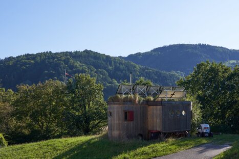 Kleines Holzhaus mit begrüntem Dach, Solarpanel und Satellitenschüssel; rundes Fenster, roter Eingang und Waldkulisse – ein Bild stiller Autarkie im Grünen.