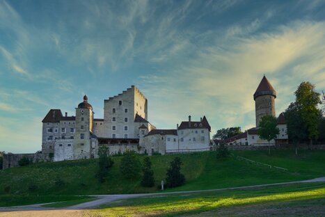 Burg Clam mit rundem Turm, Giebel und rotem Dach über grünem Hang; dramatischer Himmel und spätes Licht verleihen der Szene historische Tiefe und Bildspannung.