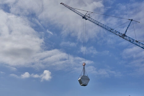 Ein großer Baukran hebt ein pentagonförmiges Metallobjekt in luftige Höhe; die Szene zeigt technische Präzision und die Dynamik moderner Infrastrukturentwicklung.