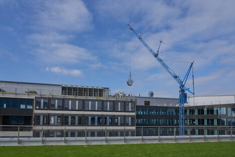 Ein blauer Baukran hebt ein sechseckiges Objekt über das neue Gebäude mit Glasfassade; die Szene verbindet technische Dynamik mit architektonischer Klarheit.