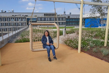 Zwischen Blumenbeeten und modernen Gebäuden lädt eine große Netzschaukel zum Verweilen ein; ein verspielter Ruhepunkt im Wandel zwischen Baukran und Freizeitgestaltung.