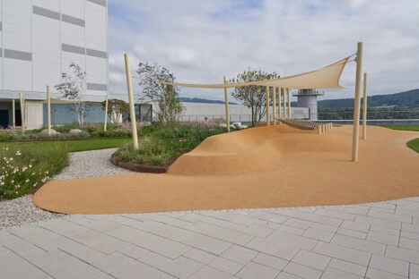 Ein orangefarbener Hügel mit Seilnetz und Schattenstruktur bildet das Zentrum des Spielbereichs; umgeben von Blumenbeeten und klarer Architektur entsteht ein sicherer, lebendiger Bewegungsraum.