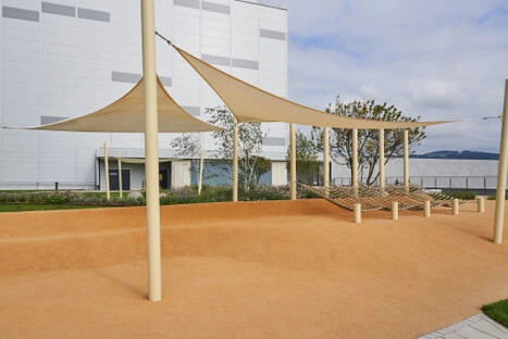 Zwischen Blumenbeeten und moderner Architektur laden gespannte Seilflächen zum entspannten Liegen ein; ein ruhiger Rückzugsort mit Blick auf Himmel und Hafen.