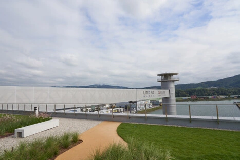 Am geschwungenen Kiesweg lädt eine weiße Bank zum Verweilen ein; zwischen Zierpflanzen und Hafeninfrastruktur entsteht ein stiller Ort mit Aussicht auf Wasser und Hügel.